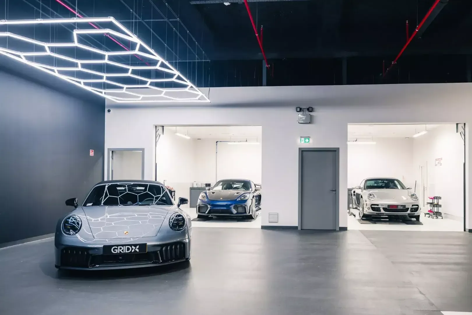 Porsche 911 grise sous les luminaires hexagonaux de l’atelier, avec deux cabines de travail à l’arrière-plan