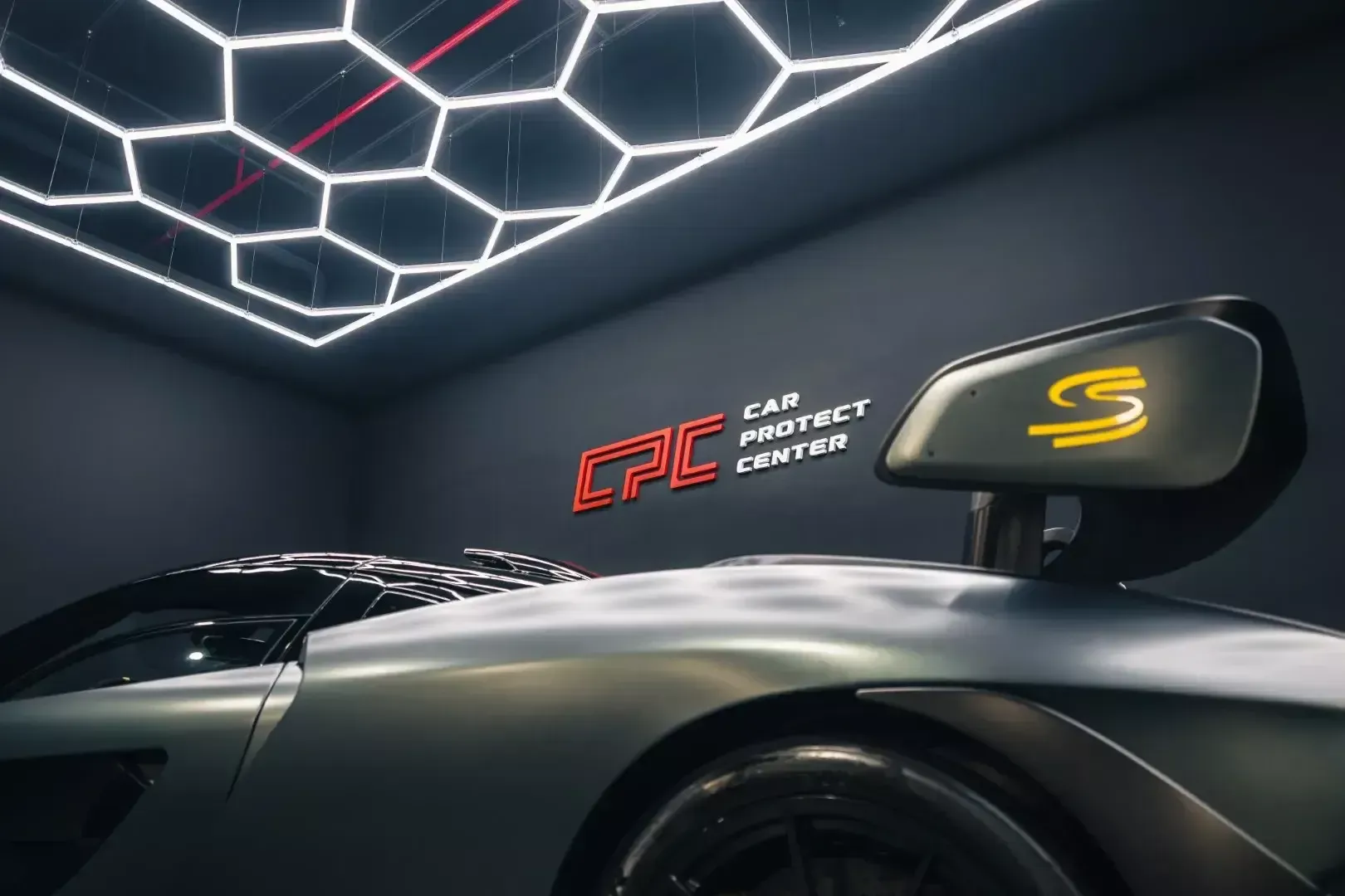 Voiture sombre sous un plafonnier hexagonal, devant le logo Car Protect Center peint sur le mur de l’atelier