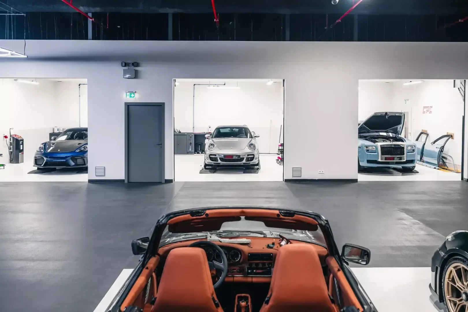 Trois cabines de travail blanches, avec une Porsche et une Rolls-Royce en cours d’intervention