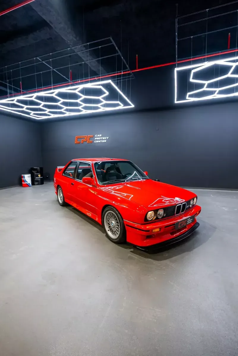 BMW M3 E30 rouge, jantes à croisillons, sous les luminaires hexagonaux de l’atelier